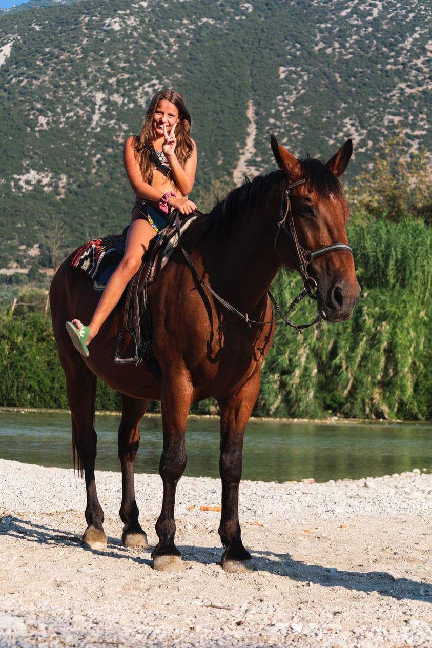A smiling woman making a peace sign while sitting on a brown horse near a body of water with mountains and greenery in the background.